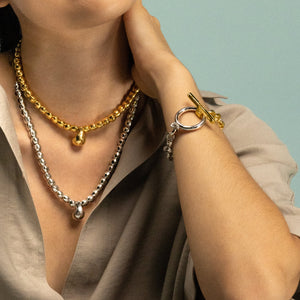 Imagen de una mujer mostrando joyería elegante. Lleva dos collares: uno superior de oro con cuentas y un colgante dorado, y otro inferior de plata con cuentas y un colgante plateado. En su muñeca se ve una pulsera que combina oro y plata con un cierre llamativo. La mujer viste una prenda de color beige claro. El fondo es de color verde suave. La imagen se enfoca en el cuello, pecho y brazo de la mujer, destacando las joyas.