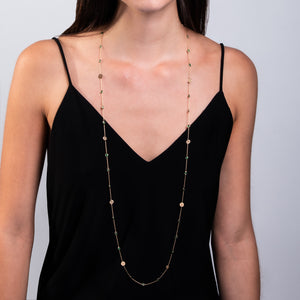 Mujer con vestido negro de tirantes luciendo un collar largo dorado. El collar es una cadena delgada con pequeñas esmeraldas y discos dorados intercalados. Cae sobre el escote en V del vestido, creando un elegante contraste con la tela oscura. La imagen se enfoca en el torso de la mujer, mostrando el collar en detalle contra su piel clara y el vestido negro.