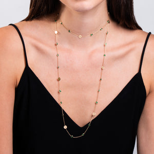 Mujer usando un collar largo de oro con pequeños discos dorados y esmeraldas, que se envuelve en doble vuelta alrededor de su cuello, creando un look elegante y sofisticado sobre un vestido negro sin mangas.