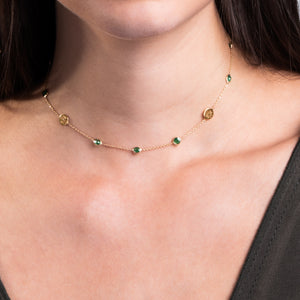 Mujer luciendo un delicado collar de oro con piedras verdes. La cadena fina alterna pequeñas piedras verdes y discos dorados con diseño de trébol. El collar descansa elegantemente sobre el cuello de la mujer, contrastando con su piel clara y cabello oscuro. La imagen se enfoca en el cuello y parte superior del pecho, mostrando el collar en detalle.